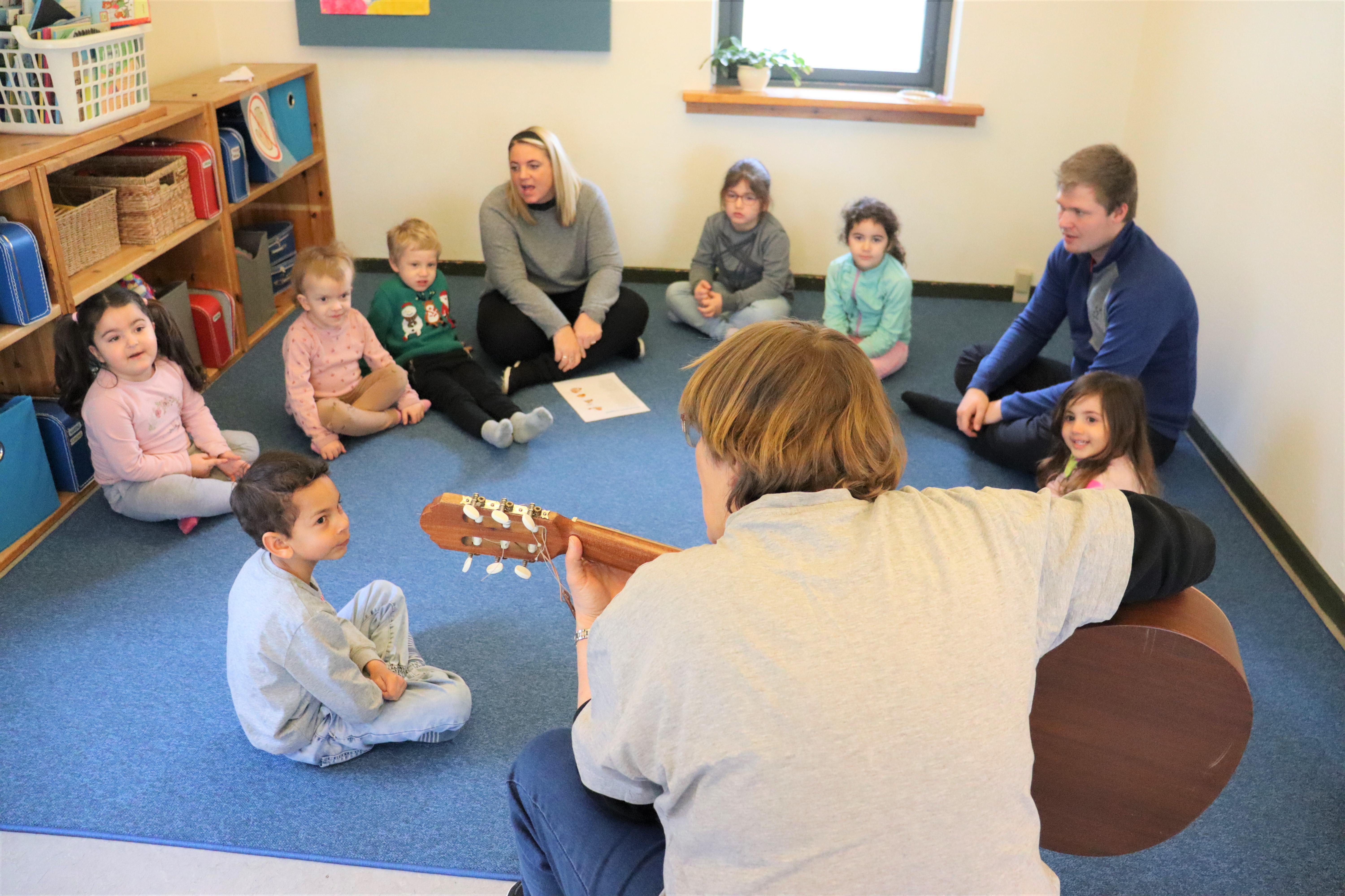 Pædagog sidder med ryggen til og spiller guitar, hvor børnene er samlet i en cirkle omkring pædagogen